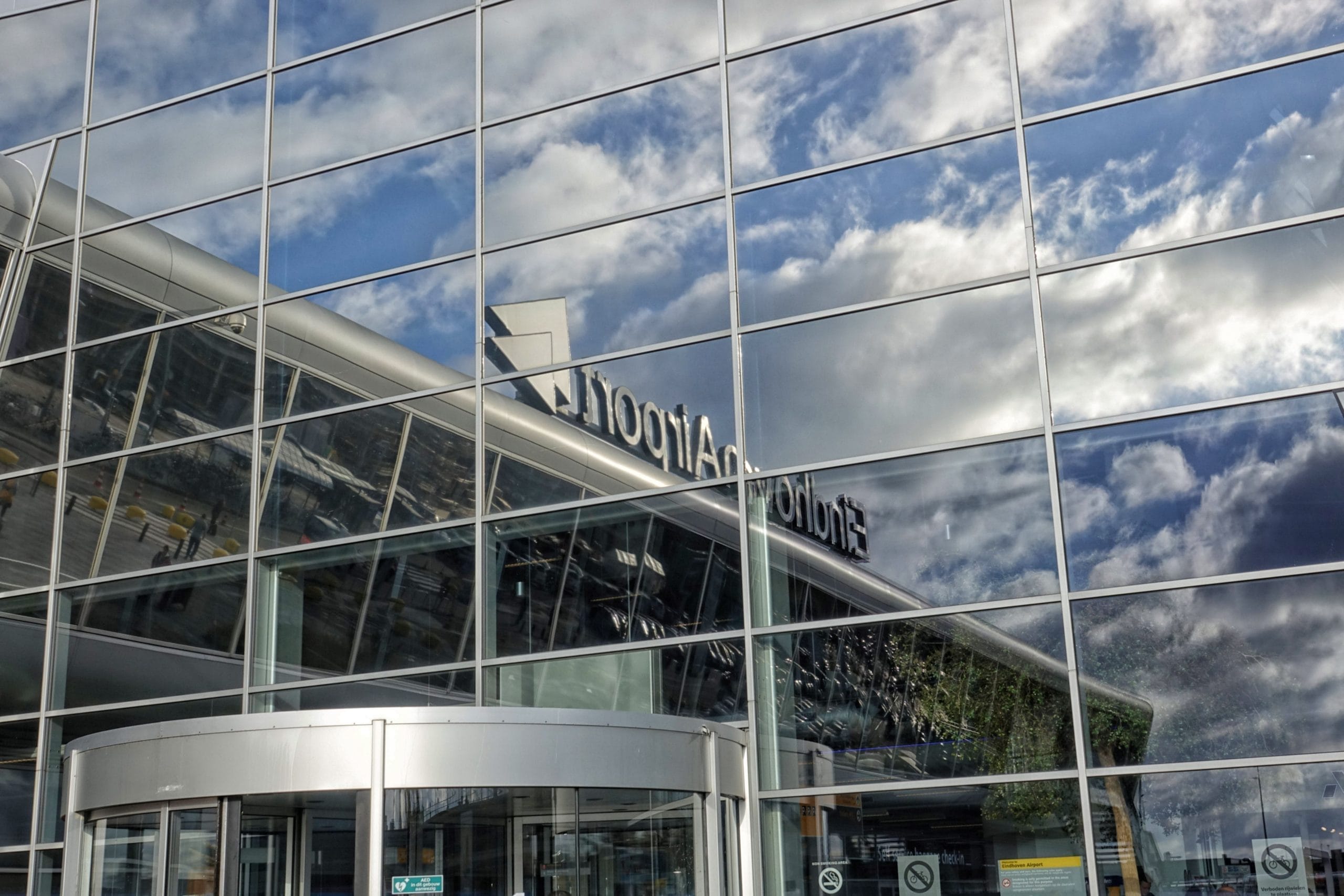 An image of Eindhoven Airport's glass front with the sign of the aiprot reflecting in the silver windows below a blue sky with white clouds.