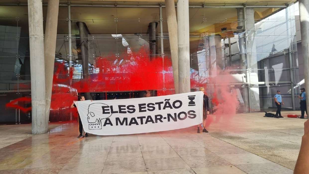 Activists holding banner saying "they are killing us" in front of the World Aviation Fest that was painted red. They claim that the aviation industry is killing us, by fueling climate breakdown