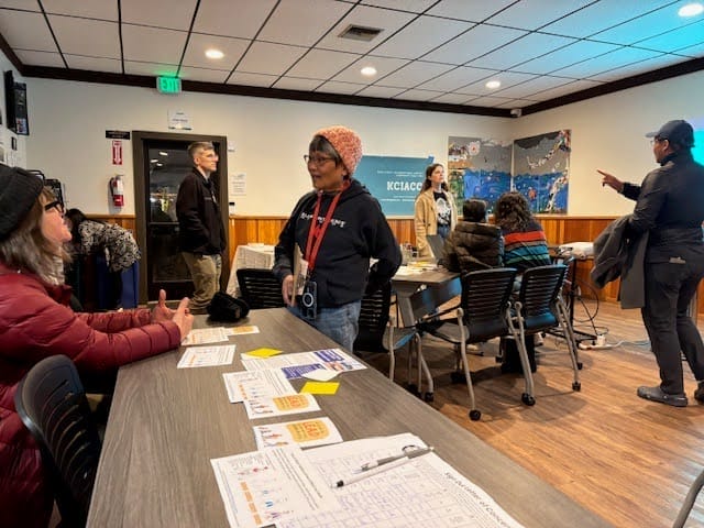 Organizadores en un acto de organización comunitaria. La foto muestra una sala con gente de pie y sentada alrededor de mesas charlando.